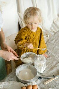 A young girl learns baking in a cozy kitchen, sifting flour with adult assistance.